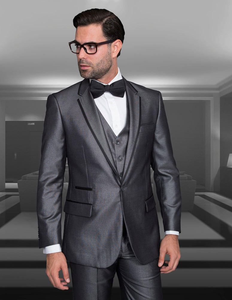 A man in a Venetian-Grey tailored fit suit from Statement Clothing, complemented by a bow tie and glasses, stands in a dimly lit room featuring modern furniture.