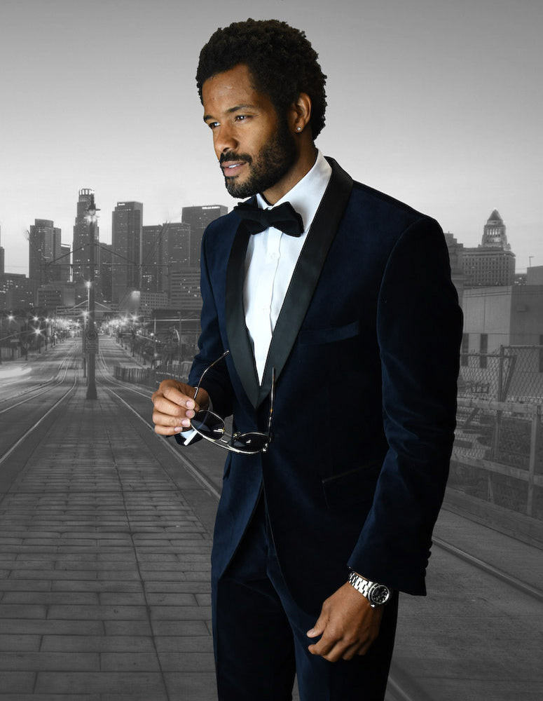 Dressed in the sophisticated STATEMENT CLOTHING | VL-100-NAVY by Statement Clothing, a man dons a tailored velvet suit with a bow tie and navy accents. He holds sunglasses while standing on an urban bridge, framed by the city skyline.