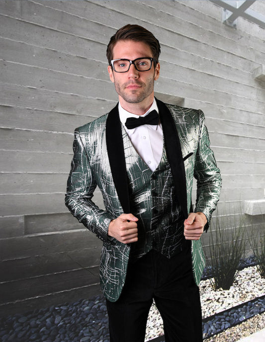 A man clad in the STATEMENT CLOTHING | VL-102-GREEN suit by Statement Clothing, featuring a shiny, textured green jacket with black lapels and tailored fit velvet pants, complemented by a bow tie, standing before a concrete wall and rocks.