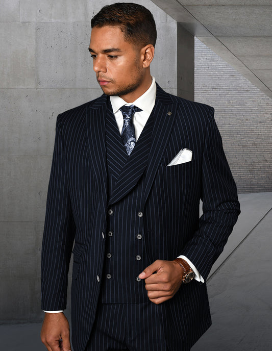 A man dressed in the ZARELLI-NAVY by Statement Clothing, featuring a pinstripe suit and tie, stands indoors against a concrete wall. His double-breasted vest subtly complements his ensemble as he gazes to the side.