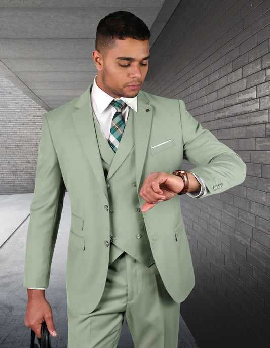 A man wearing the STATEMENT CLOTHING | ZARGALA-APPLEGREEN suit from Statement Clothing, paired with a patterned tie, checks his watch while standing in a corridor with brick walls.