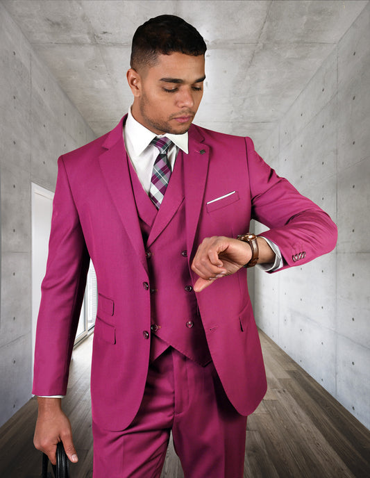 A man in a vibrant ZARGALA-FUCHSIA suit by Statement Clothing checks his watch while standing in a sleek, modern gray hallway.