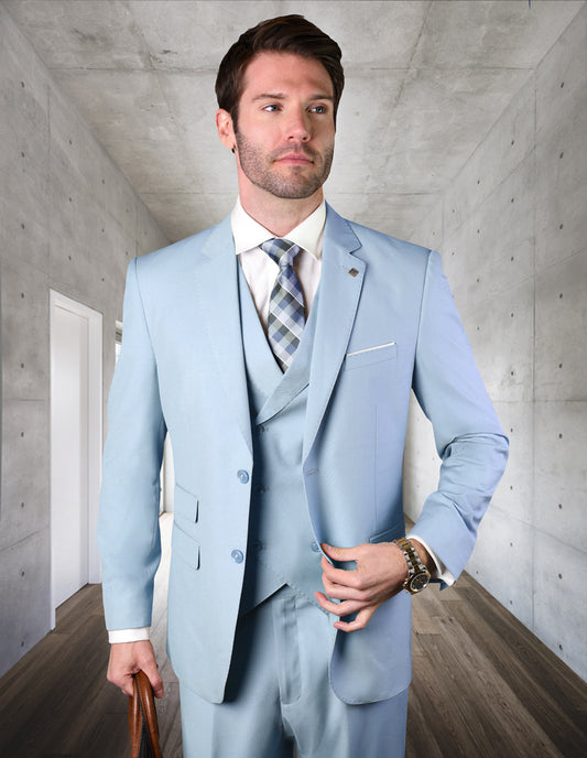 A man dressed in the ZARGALA-POWDERBLUE suit from Statement Clothing, paired with a checkered tie, stands in a modern hallway. He showcases the elegance of Italian wool against the backdrop of concrete walls and wooden flooring.