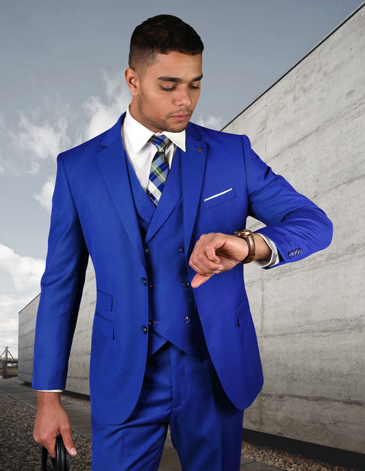 A man wearing the STATEMENT CLOTHING | ZARGALA-ROYALBLUE suit from Statement Clothing, expertly tailored with a modern fit and crafted from fine Italian wool, checks his watch while standing outdoors against a concrete wall backdrop.