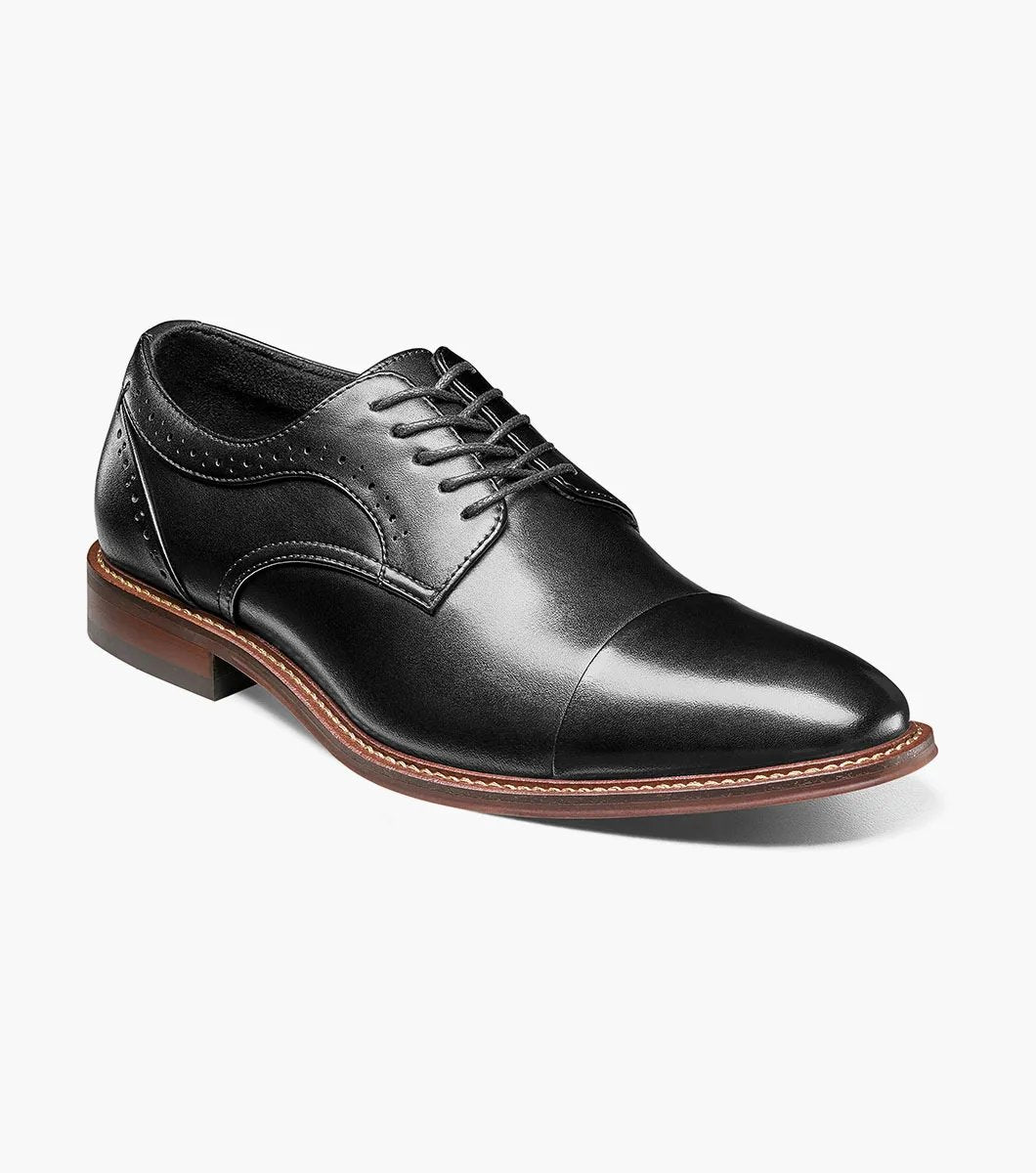 The Stacy Adams - MADDOX Cap Toe Oxford shoe features a black smooth leather finish with a polished appearance, complemented by a brown sole, intricate decorative stitching, and anatomical arch support.