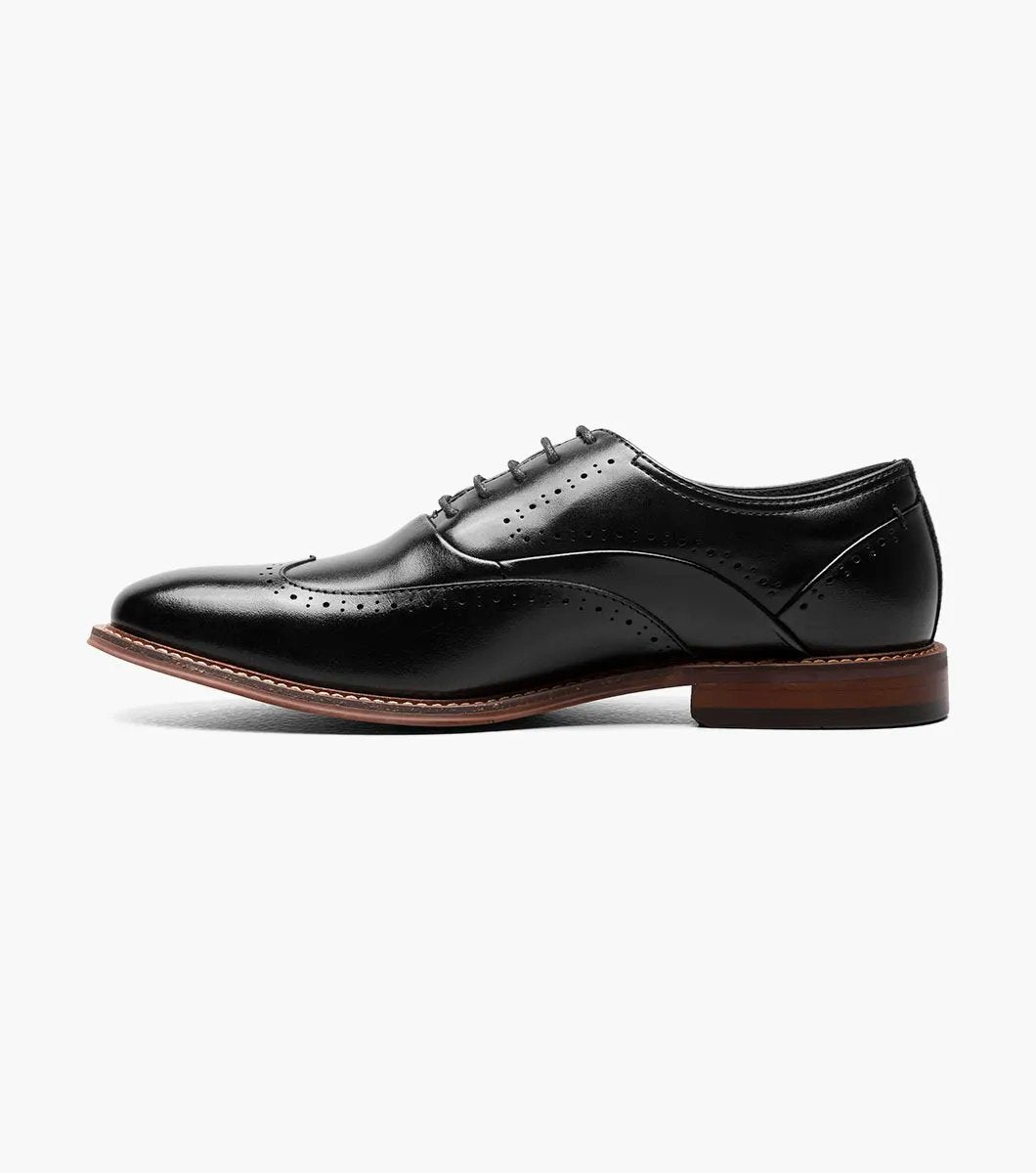 The Stacy Adams MACARTHUR Wingtip Oxford in Black Smooth, featuring decorative perforations and a sophisticated wingtip design with a brown heel and lace-up closure, is elegantly displayed on a white background.