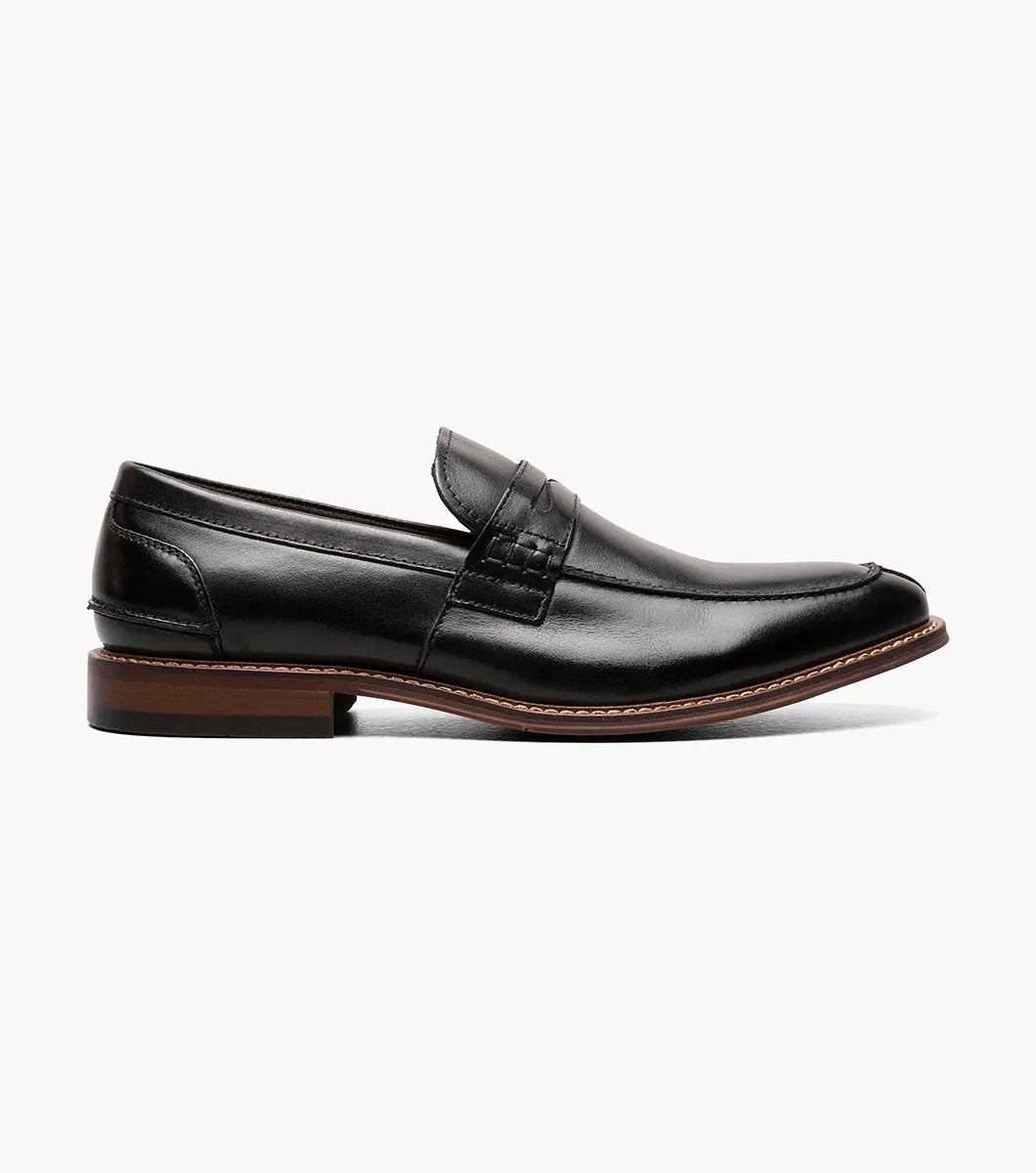 The Stacy Adams MARLOWE Algonquin Moc Toe Penny Slip On in black, featuring a refined leather upper and a contrasting brown sole, is displayed against a plain background.