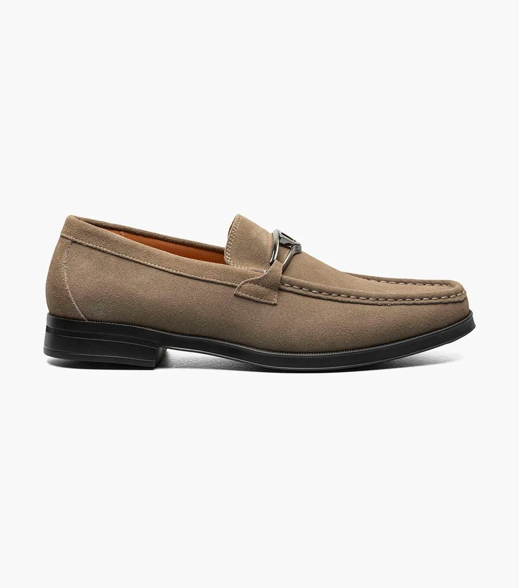 The Stacy Adams PALLADIAN Moc Toe Slip On in Fossil Suede features a brown suede leather design with a black sole, adorned with a silver buckle detail on top, and enhanced with memory foam for ultimate comfort.