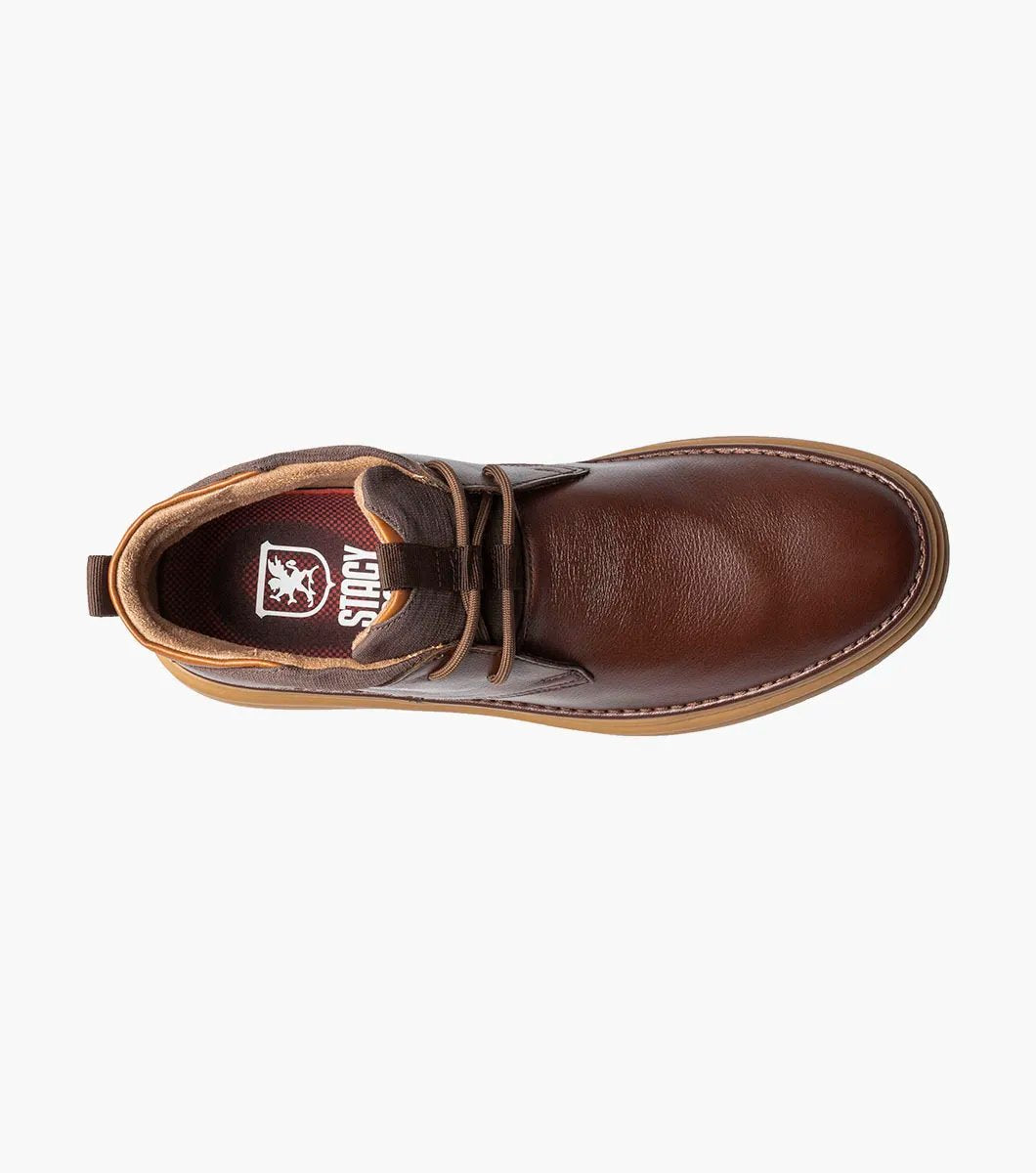 The Stacy Adams DELSON Plain Toe Chukka Boot in Chocolate, style number 25556-202, features a brown leather design with a rounded toe, lace-up front, and a cushioned brown sole with anatomical arch support, all presented against a white background.