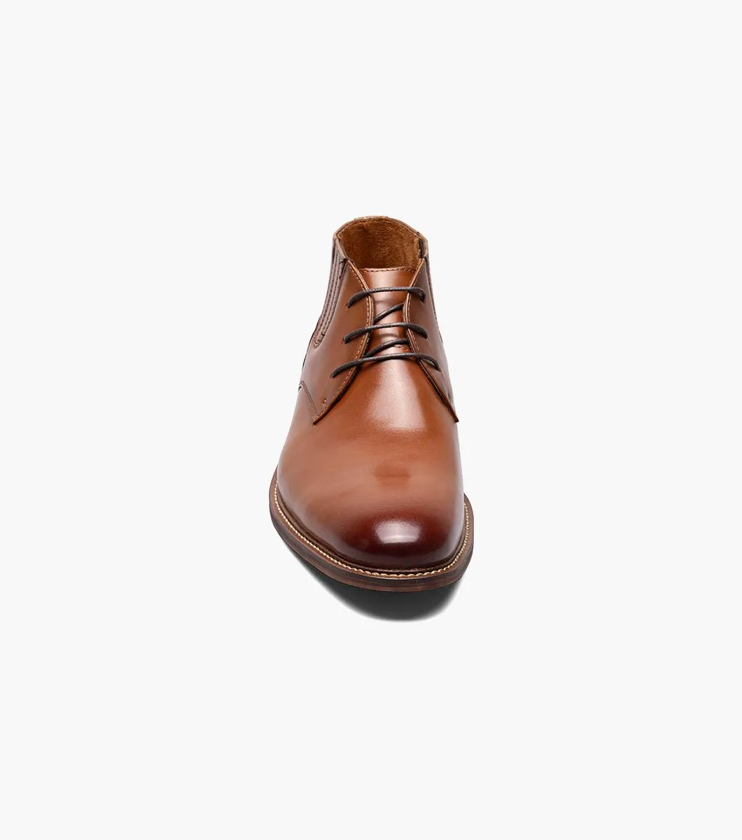 The Stacy Adams Maxwell Plain Toe Chukka Boot in Cognac features a sophisticated leather design with three eyelets, detailed stitching, and a versatile low heel, making it ideal for both dressy and casual events.