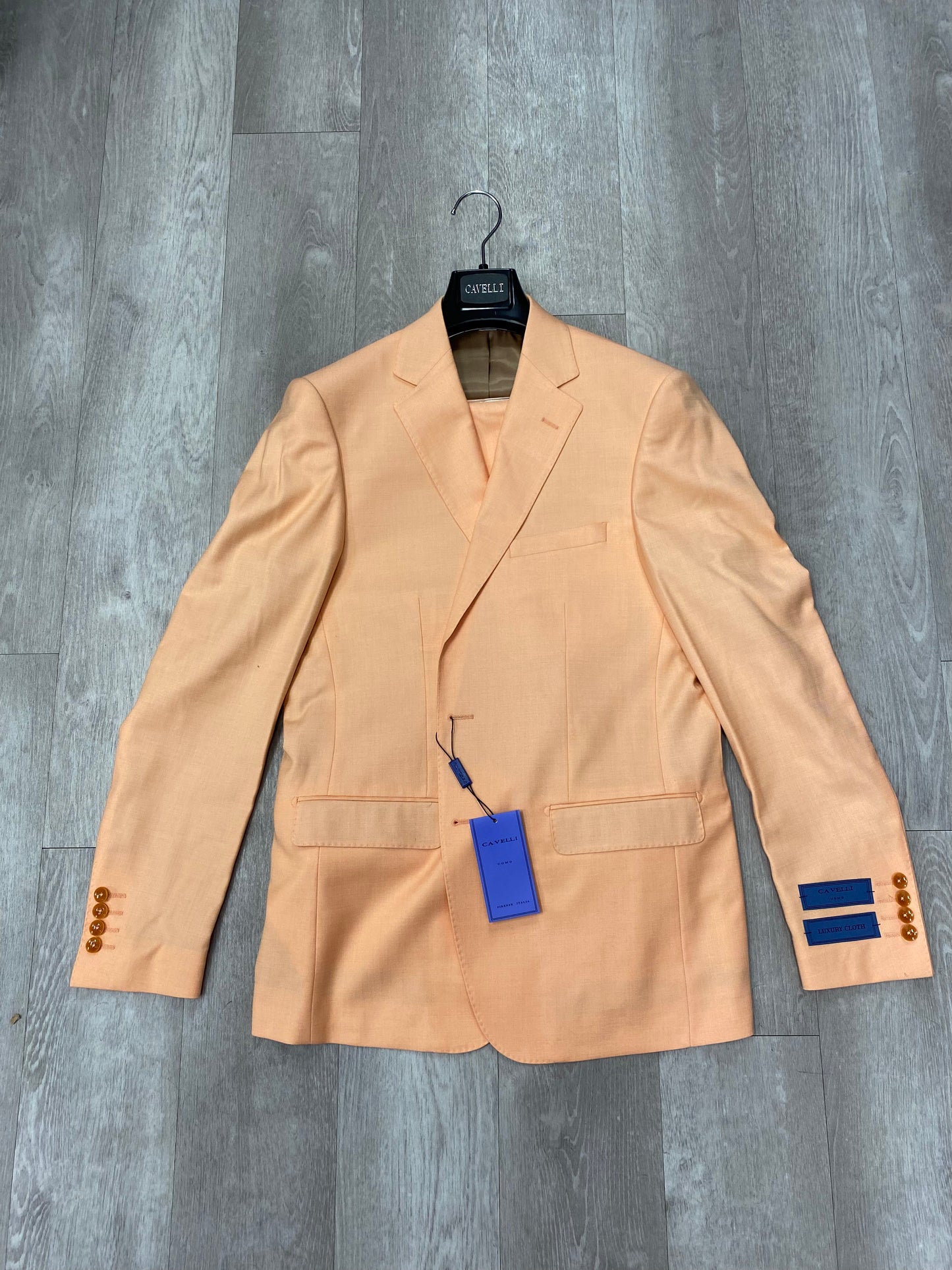 The Cavelli Uomo Porto Slim Fit Suit 1986/11 Light Orange, from the CAVELLI brand, elegantly hangs with its tag and label on a hanger against a wooden floor backdrop. It features brown buttons and offers lightweight comfort suitable for any occasion.