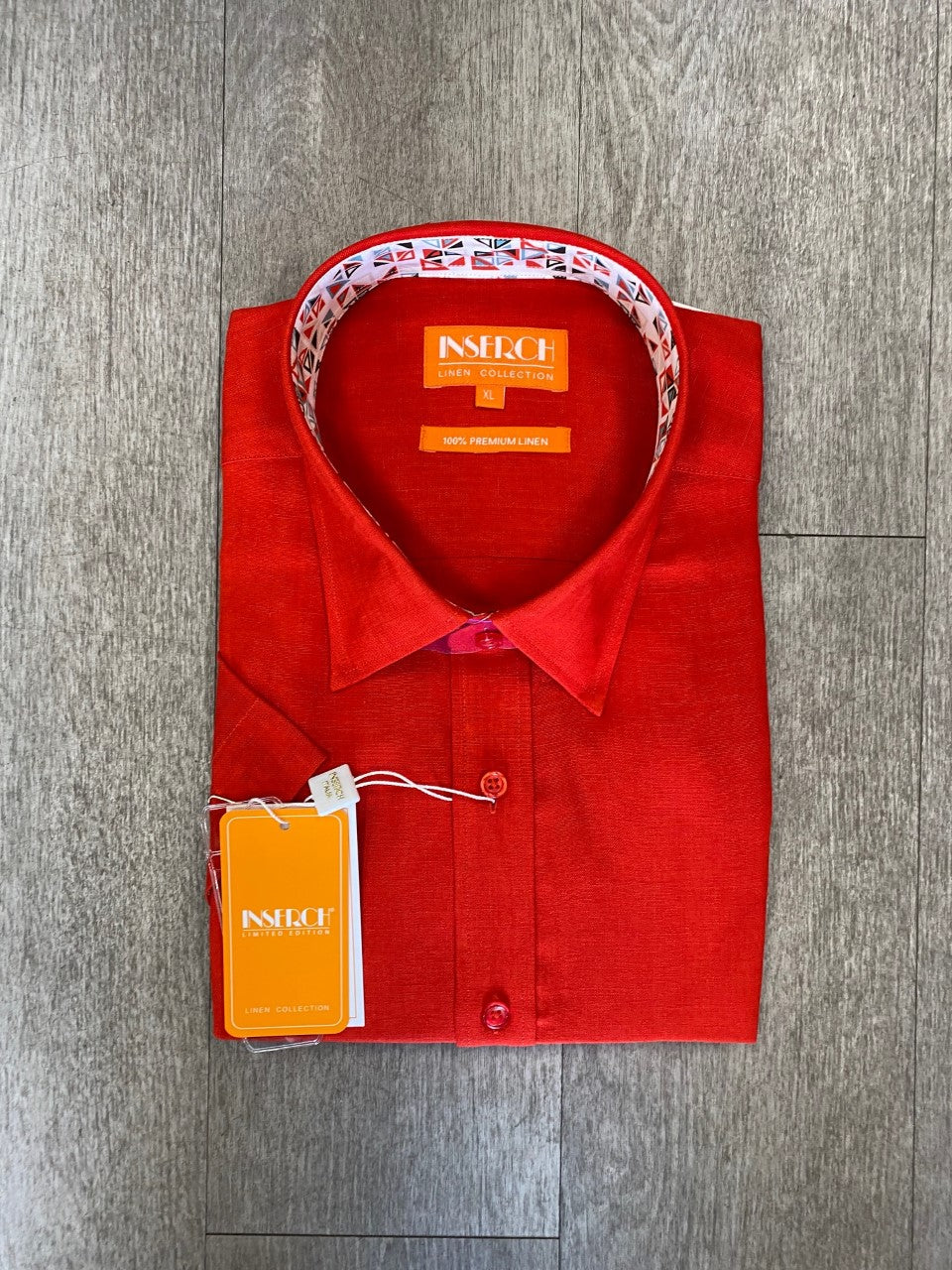 A folded Inserch Premium Linen Yarn Dye Short Sleeve Shirt in red, featuring a printed inner collar and brand tags, is displayed on a wooden surface.