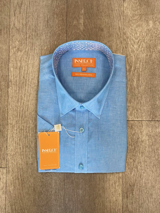 A folded Premium Linen Yarn Dye Short Sleeve Shirt in light blue, featuring a patterned collar lining, is placed on a wooden surface with Inserch brand tags attached.